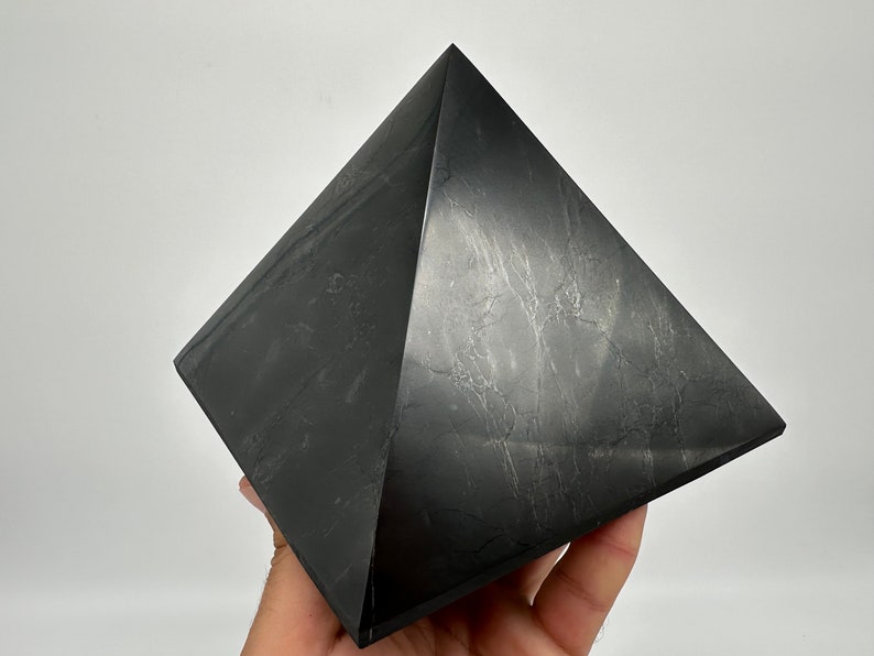 May include: A black obsidian pyramid crystal. The pyramid is smooth and polished, with a slight sheen. It is a dark gray color with subtle white veins running through it.