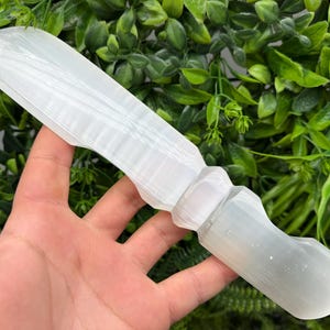 Puede incluir: Una varita de selenita blanca, sobre un fondo de follaje verde. La varita tiene una forma lisa y alargada con una base en forma de mango y una punta redondeada. El cristal es translúcido con estrías visibles.