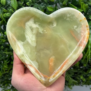 Op de afbeelding: Een hartvormige, groene onyx schaal met oranje en witte adering. De schaal wordt in een hand gehouden, met een achtergrond van groen gebladerte. De onyx schaal is een decoratief item.