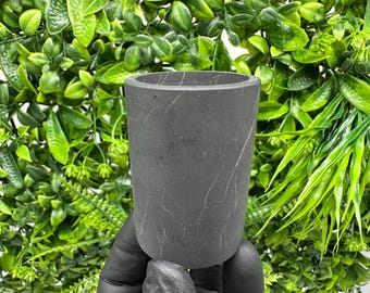 May include: A cylindrical, dark gray container with a marble-like pattern. The container is held against a backdrop of lush green foliage. The vessel has a smooth, matte finish and a simple, modern design. Ideal for holding small items or as a decorative piece.