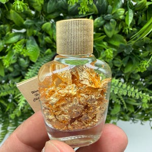 May include: A small, clear glass bottle filled with gold flakes, topped with a gold-colored cap. The bottle is held in front of a backdrop of green foliage. A small tag with text is visible.