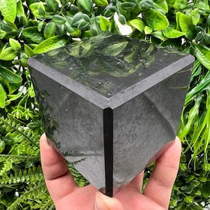 Könnte beinhalten: Ein dunkelgrauer, geometrischer Kristall, möglicherweise ein Dekorationsartikel oder ein Briefbeschwerer. Der Kristall hat ein facettiertes Design und eine polierte Oberfläche. Der Hintergrund zeigt üppiges grünes Laub, das einen Kontrast zum dunklen Kristall bildet.