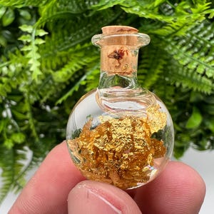 May include: A small, round glass bottle with a cork stopper filled with gold leaf flakes. The bottle is clear and the gold leaf is visible inside.