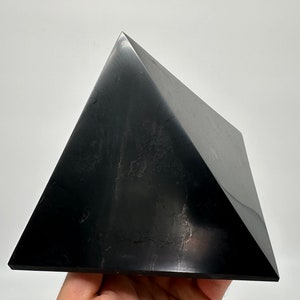 May include: A black obsidian pyramid crystal. The pyramid is smooth and polished, with a slight sheen. It is a dark, opaque black color.