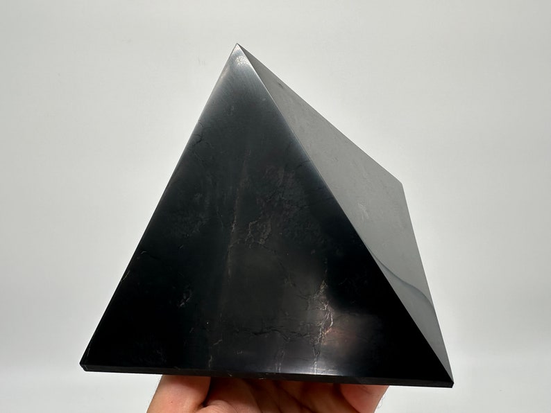 May include: A black obsidian pyramid crystal. The pyramid is smooth and polished, with a slight sheen. It is a dark, opaque black color.