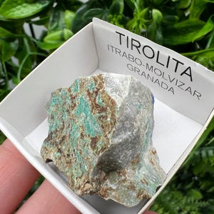 May include: A green and gray Tirolite mineral specimen in a white box with the label "TIROLITA ITRABO-MOLVIZAR GRANADA".