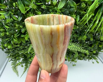 May include: A tapered drinking cup carved from polished banded onyx stone, featuring a smooth surface with natural vertical striations in shades of pale green, cream, amber, and reddish-brown, showcasing the unique mineral layers of the material.