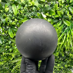 Può includere: Una sfera nera opaca, tenuta da una mano guantata, su uno sfondo di vivace fogliame verde. La superficie della sfera presenta sottili imperfezioni, che aggiungono alla sua consistenza unica. L'estetica generale è moderna e minimalista.