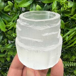 Può includere: Un portacandele cilindrico in cristallo di selenite trasparente. Il supporto ha un design a più livelli con una base ampia e un'apertura circolare nella parte superiore. La selenite ha un aspetto traslucido e cristallino. Lo sfondo è un fogliame verde sfocato.