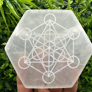 Può includere: Piatto di ricarica in selenite bianca con un simbolo di fiore della vita inciso. Il piatto è esagonale e ha una superficie liscia e lucida.