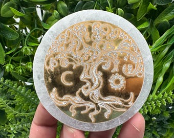 May include: A white circular stone coaster with a gold tree of life design. The tree has intricate details and is surrounded by a crescent moon and a sun.