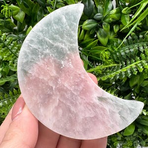 Può includere: Un cristallo di selenite a forma di luna crescente di colore rosa e bianco. Il cristallo ha una superficie ruvida e testurizzata.