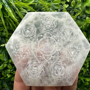 May include: White selenite charging plate with seven chakra symbols etched into the surface. The plate is hexagonal in shape and has a smooth, polished finish.