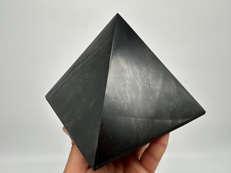 May include: A black obsidian pyramid crystal. The pyramid is smooth and polished with a slight sheen. The crystal is a dark gray color with subtle white veins.