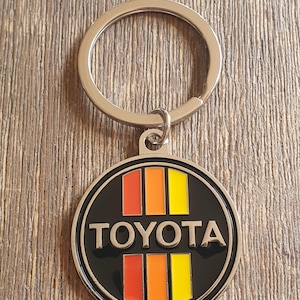 May include: A silver key ring with a round black and silver charm. The charm features a red, orange and yellow striped design with the word "TOYOTA" in white lettering.