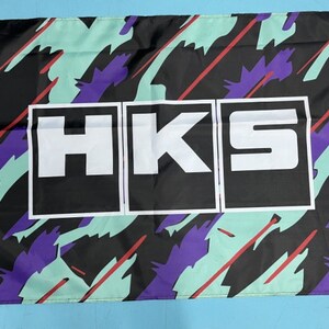 May include: A black and white flag with a colourful abstract pattern. The flag features the word "HKS" in white letters on a black background.