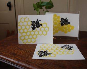 Puede incluir: Tres tarjetas blancas con un patrón de panal amarillo e ilustraciones de abejas negras. Las tarjetas se exhiben sobre una superficie de madera, con una planta en el fondo. El diseño de la abeja se repite en cada tarjeta.