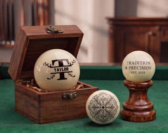 May include: Three off-white billiard balls with black designs and a wooden box. One ball features the letter T and the name TAYLOR, another displays the text TRADITION & PRECISION EST. 1920, and the third shows a decorative floral pattern.