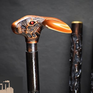 Puede incluir: Un bastón de madera con un mango tallado en forma de cabeza de conejo. Los ojos del conejo son rojos y el mango es de color cobre. El bastón es de color marrón oscuro y tiene un diseño tallado.