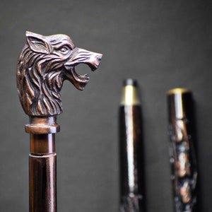 Puede incluir: Un bastón de madera con un mango de cabeza de lobo. El bastón es de color marrón oscuro y tiene tallas intrincadas. Se pueden ver otros dos bastones en el fondo.