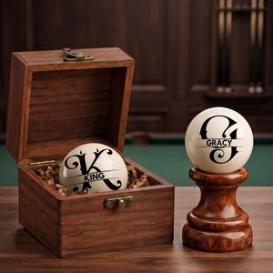 May include: A dark wood box with a metal latch containing a white billiard ball personalized with a black letter K and the name KING. Beside it, a second white billiard ball with a black letter G and the name GRACY rests on a wood stand.