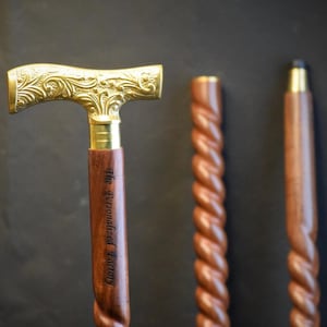 Puede incluir: Tres bastones de madera para caminar. Uno tiene una parte superior adornada de color dorado con el texto "The Personalized Factory" grabado en el eje. Los otros dos bastones tienen un diseño retorcido.