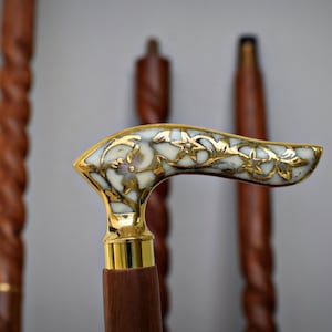 Puede incluir: Un bastón con un mango adornado en blanco y dorado. El mango tiene forma de gancho curvo y presenta un diseño floral.