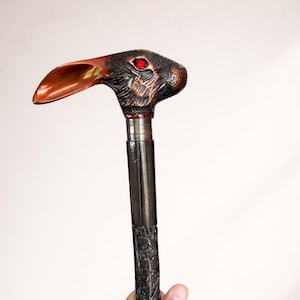 May include: A walking stick featuring a handle shaped like a rabbit head with a metallic copper finish, one prominent ear, and a bright red eye. The shaft is dark, textured, and carved with intricate patterns along its entire length.