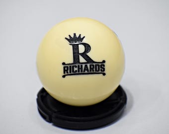 May include: A white billiard ball with a black logo featuring a crown and the letter "R" above the word "RICHARDS".