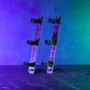 Könnte beinhalten: Zwei vertikale schwarze und violette Gaming-Headset-Ständer. Die Ständer haben ein geometrisches Muster und drei schwarze Stützarme. Die Ständer stehen auf einer dunklen Oberfläche mit einem lila-grünen Farbverlauf im Hintergrund.