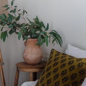 Könnte beinhalten: Eine Terrakotta-Vase mit grünen Zweigen steht auf einem kleinen Holzschemel. Ein senffarbenes Kissen mit dunklem Blumenmuster liegt auf einem weißen Bett. Die Szene strahlt eine gemütliche, natürliche Ästhetik aus.
