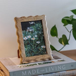 May include: A light-brown wooden picture frame with a wavy edge displays a photo of white daisies. The frame rests on two books, one titled "MADE FOR LIVING." The scene is set on a wooden surface, with a green plant in the background.