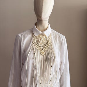 Puede incluir: Una camisa blanca de manga larga con botones y una pieza de cuello de macramé decorativa. El diseño de cordón color hueso incluye patrones de nudos intrincados y flecos colgantes acentuados con pequeñas cuentas de metal plateado.