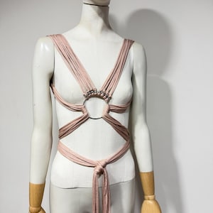 Puede incluir: Un top cruzado de color rosa empolvado con múltiples tiras de tela finas que convergen en un anillo central de metal plateado. El diseño incluye pequeñas cuentas plateadas en las tiras superiores y se ata a la cintura con un nudo.