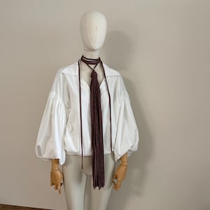 Peut inclure: Un chemisier blanc à manches longues avec un col pointu et des manches bouffantes froncées, associé à un long collier cordon marron foncé doté d'un nœud central et d'une frange en cascade composée de fines bandes de tissu pendantes.