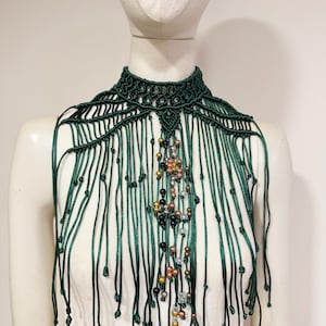 May include: A dark green macrame choker necklace featuring an intricate knotted collar design with long, dangling fringe cords. The center of the piece is embellished with a vertical arrangement of metallic, black, and clear beads.