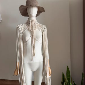 Può includere: Un coprispalle in macramè beige con lunghe maniche a frange e un laccetto al collo, indossato sopra un top bianco semplice su un manichino. Il manichino indossa anche un cappello marrone testurizzato a tesa larga. Il macramè presenta nodi complessi.