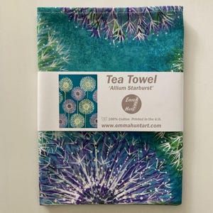 Puede incluir: Paño de cocina doblado con un fondo turquesa y un gran diseño de diente de león morado y blanco. La toalla tiene una banda blanca con el texto "Tea Towel 'Allium Starburst'" y el nombre de la marca. 100% algodón.