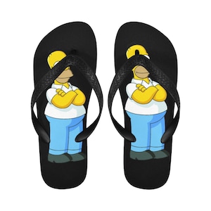 Puede incluir: Un par de chanclas negras con un personaje de dibujos animados amarillo que lleva pantalones vaqueros azules y una camisa blanca. El personaje tiene una cara amarilla y una gran sonrisa.