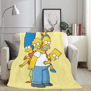 Op de afbeelding: Gele deken met de Simpsons-familie, Homer, Marge, Bart en Lisa, in cartoon-stijl. De deken is geel met een witte rand.