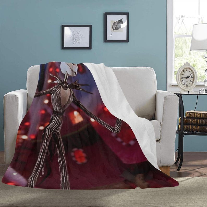 May include: A red, white, and black throw blanket with a printed image of Jack Skellington from the Nightmare Before Christmas movie. The blanket is folded and draped over a white couch.