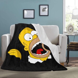 Op de afbeelding: Een zwart-gele deken met een cartoon afbeelding van Homer Simpson uit The Simpsons. Homer wordt afgebeeld met zijn mond open en zijn tong uitstekend. De deken is gemaakt van zacht, pluche materiaal.