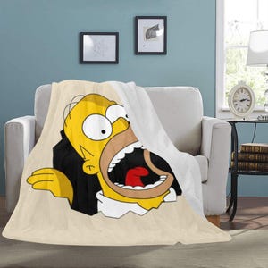 Op de afbeelding: Een beige fleece deken met een grote cartoonafbeelding van Homer Simpson met zijn mond wijd open in een schreeuw. De deken is over een witte fauteuil gedrapeerd, met een deel van de witte achterkant zichtbaar.