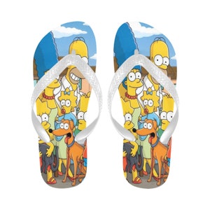 Puede incluir: Un par de chanclas amarillas y azules con una tira blanca. Las chanclas presentan un estampado de la familia Simpson, incluyendo a Homer, Marge, Bart, Lisa, Maggie, el pequeño ayudante de Santa y Bola de Nieve.