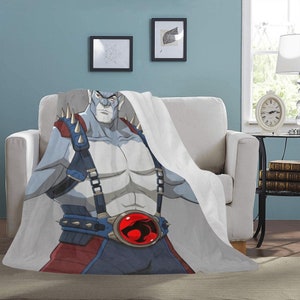 Może przedstawiać: Biały koc z polaru z kreskówkowym wizerunkiem niebieskoskórego bohatera w czerwono-niebiesko-czarnym stroju. Bohater ma czerwony symbol na klatce piersiowej z białym tygrysem w środku.