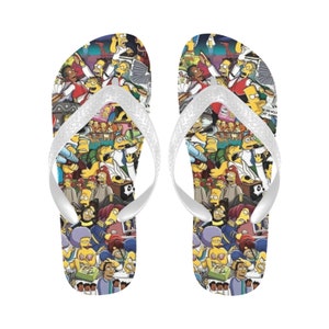 Puede incluir: Un par de chanclas blancas con un estampado colorido de personajes de la caricatura Los Simpson. El estampado presenta una variedad de personajes, incluyendo a Homero, Marge, Bart, Lisa y Maggie.