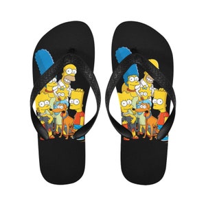 Puede incluir: Un par de chanclas negras con una imagen colorida de la familia Simpson en la parte delantera. La imagen incluye a Homer, Marge, Bart, Lisa, Maggie y el perro de la familia, Santa's Little Helper.