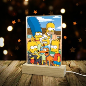 May include: A rectangular, illuminated night light featuring the Simpson family. The image shows Homer, Marge, Bart, Lisa, Maggie, and their pets. The light sits on a wooden base with a white cord.