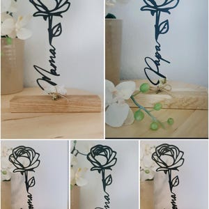 May include: Black laser-cut wooden rose with a stem and the word "Mama" in cursive script. The rose is mounted on a small wooden block. The background includes white flowers and a neutral-toned surface.