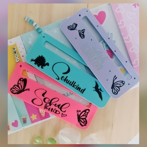 Puede incluir: Tres marcapáginas coloridos con rectángulos recortados. El marcapáginas rosa tiene diseños de mariposas y la palabra "Schulkind". El marcapáginas turquesa tiene un dinosaurio y la palabra "Schulkind". El marcapáginas morado tiene mariposas y la palabra "Schulkind".